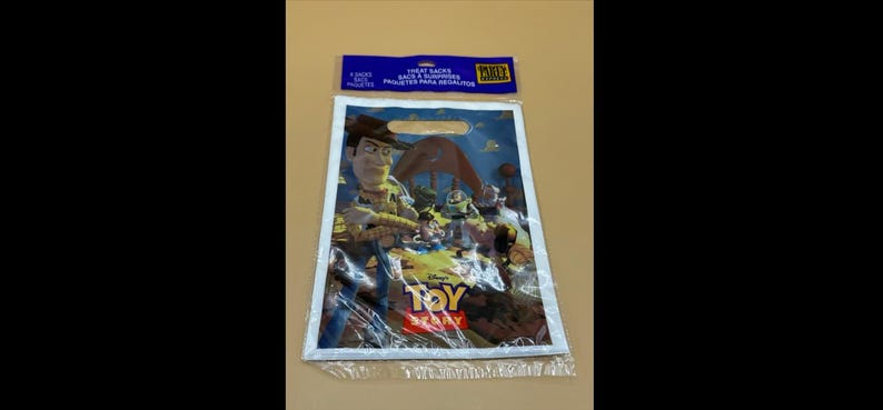 May include: A clear plastic party favor bag featuring characters from the animated film "Toy Story." The bag has a white border and a blue header with text. The bag's design includes Woody, Buzz Lightyear, and the Toy Story logo.