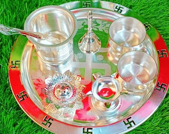 May include: A silver tray with a red and green border, featuring a swastika design. The tray contains a variety of silver items, including a small bell, a cup, a bowl, a small incense burner, and a small oil lamp. A silver matchbox is sitting on the green grass.