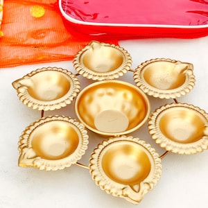 May include: A golden metal diya set, arranged in a circular pattern. The set includes seven small, ornate bowls with scalloped edges, designed to hold oil and a wick for traditional lighting. A red bag is visible in the background.