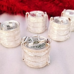 May include: A collection of silver-coloured, tiered containers with handles. Each container features a detailed floral pattern. The containers are stacked and resemble lunch boxes or food carriers. The background is a soft red and white.