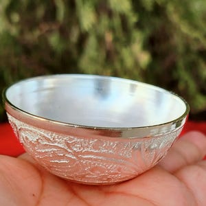 May include: A small, silver-coloured bowl with an embossed floral design around the exterior. The bowl has a polished, reflective interior and a smooth, shiny rim. The bowl is held in a person's hand.
