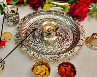 May include: A silver-colored puja thali with intricate embossed designs, holding a small oil lamp, incense, and small bowls of nuts. Red roses provide a vibrant backdrop. The thali is surrounded by other religious items.