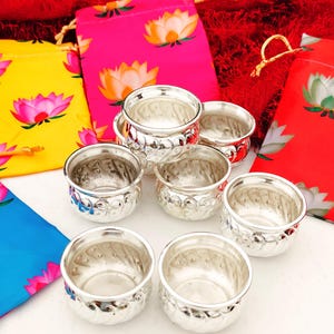 May include: A collection of small, silver-colored bowls with a decorative pattern around the rim. The bowls are arranged with colorful fabric pouches featuring lotus flower designs in pink, yellow, and red.