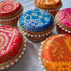 May include: Six round decorative boxes with intricate patterns and gold trim. The boxes are covered in colourful fabrics with floral and geometric designs. Each box has a row of white pearls around the edge.