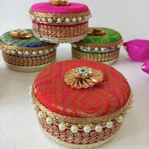 May include: Four round decorative boxes with lids. The boxes are covered in colorful fabric with gold trim and pearl accents. The lids have a jeweled flower design.