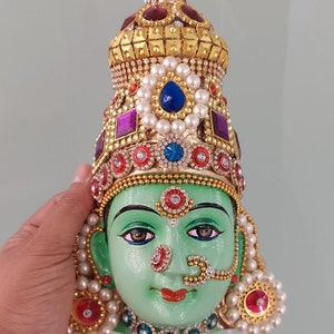 May include: A green face mask adorned with elaborate gold and jewel embellishments. The mask features a crown, earrings, and a nose ring. The face is painted with detailed features and a serene expression.