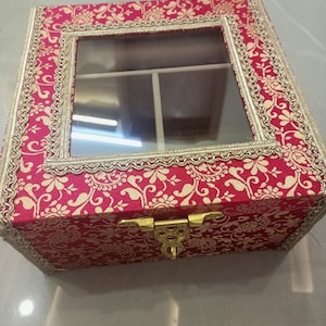 May include: A square storage box featuring a vibrant magenta fabric exterior with a gold floral pattern. The lid includes a clear glass window framed by decorative gold trim. A gold-toned metal latch secures the front of the box.