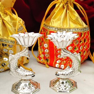 May include: Two silver-colored decorative candle holders, each featuring a fish design supporting a flower-shaped bowl. The holders sit on a hexagonal base. Gold and red drawstring bags are in the background.