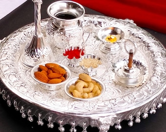 May include: A silver tray with a decorative design features a silver pitcher, a small bowl with red powder, a bowl with almonds, a bowl with cashews, a small silver bowl with rice, a silver diya with a lit candle, and a silver incense burner with a stick of incense.
