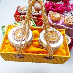 May include: A yellow and gold gift basket containing two silver containers with gold lids. The containers are decorated with gold designs and are held together by a string of white beads.