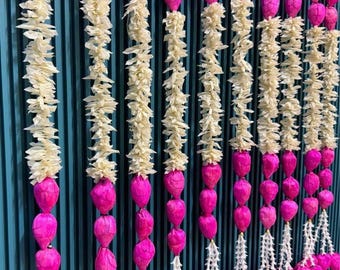 May include: A series of vertical decorative garlands featuring alternating sections of bright pink lotus buds and cream-colored ruffled flower petals. Each strand is finished with a cluster of pink buds and small white floral accents at the bottom.