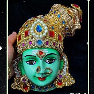 May include: A green face mask with intricate gold and jewel-toned embellishments. The mask features a traditional Indian design with a third eye and a nose ring.