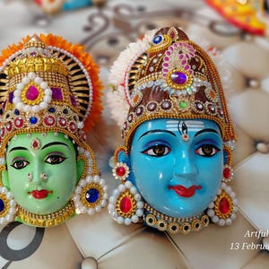 May include: Two decorative face masks, one green and one blue, adorned with ornate headwear and jewelry. The green mask has orange and gold accents, while the blue mask features brown and purple details. Both have detailed facial features.