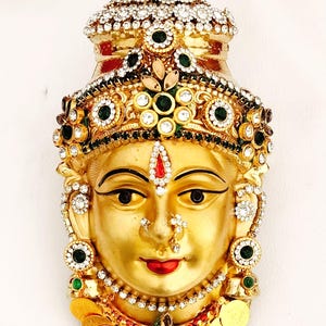May include: A gold-coloured decorative face with intricate details. The face is adorned with a jewelled crown, earrings, and a necklace. The jewels include clear, green, and red stones. The lips are painted red, and the eyes are outlined in black.