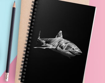May include: A black and white photo of a shark swimming in the ocean. The photo is printed on a black notebook with a spiral binding. The notebook is on a brown surface with a pink and blue background. There is a laptop, a cup of coffee, a pencil, and a pair of glasses on the surface.