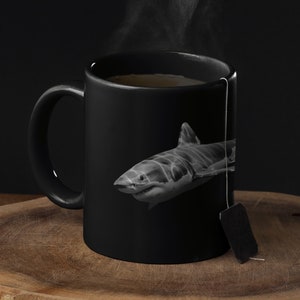 May include: Black ceramic mug featuring a grayscale image of a shark. The mug contains a hot beverage with steam rising. A tea bag is attached to the mug. The mug is placed on a wooden surface against a dark background.