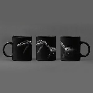 May include: Three black ceramic mugs with a white dinosaur skeleton graphic printed on each side. The dinosaur is facing right on the left mug, facing left on the right mug, and facing forward on the middle mug.