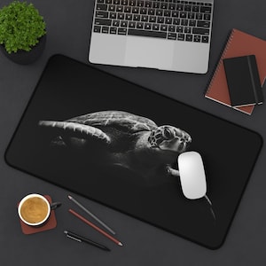 May include: Black mouse mat with a monochrome image of a sea turtle. A laptop, mouse, and stationery are in the background. The rectangular mat has a black border. The turtle is the central focus of the image.