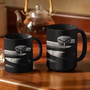 Peut inclure: Deux tasses à café noires avec une image en noir et blanc d'un serpent avec sa langue sortie. Le serpent est enroulé autour de la tasse. Les tasses sont sur une table en bois.