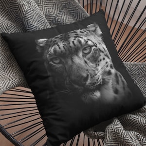 May include: Black and white close-up photograph of a snow leopard's face. The snow leopard is looking directly at the camera with a serious expression. The image is printed on a black throw pillow.