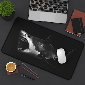 May include: Black and white mouse pad with a graphic of a shark with its mouth open. The shark is in the center of the mouse pad and is facing the right side of the image. There is a white computer mouse on the mouse pad.