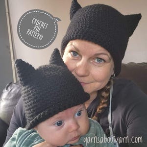 May include: A woman and a baby are wearing black crocheted hats with ears. The hats are shaped like a cat's ears. The woman is holding the baby in her arms. The text "CROCHET PDF PATTERN" is in a speech bubble in the top left corner of the image.
