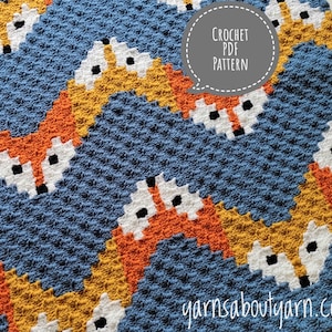 Puede incluir: Una manta de crochet con un patrón repetido de zorros naranja, blanco y negro sobre un fondo azul. El texto "CROCHET PDF PATTERN" está en un bocadillo de diálogo sobre la manta. El sitio web "yarnsaboutyarn.com" está en la esquina inferior derecha de la imagen.