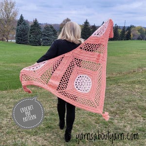 May include: A pink and white crochet shawl with a geometric pattern. The shawl is being worn by a person in a black shirt and black pants. The text "CROCHET PDF PATTERN" is in a circle on the left side of the image. The text "yarnsaboutyarn.com" is at the bottom of the image.