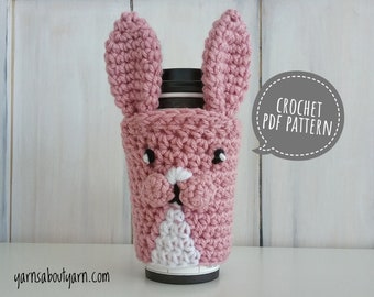 May include: Pink crocheted bunny coffee cup cozy with black eyes and a white nose. The cozy is on a white surface with a speech bubble that says "CROCHET PDF PATTERN!"