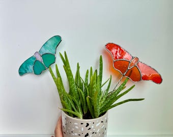 May include: Two stained glass butterfly plant stakes, one green and one orange, are placed in a potted aloe vera plant. The plant is in a speckled gray pot.