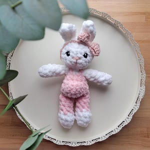 May include: A handmade crochet bunny doll with white and pink details. The bunny has a white head, ears, and arms, and is wearing a pink outfit with a matching bow. The doll is on a white decorative tray.