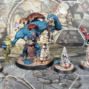 May include: A blue and green miniature fantasy creature with multiple mushrooms growing on its head, holding a green frog in its hand. The creature is standing on a base with a brown and blue painted surface. There are three other miniature fantasy creatures on bases, one with a red and black head, one with a white and gray head, and one with a green and purple head.