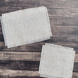 May include: Two beige burlap coasters with frayed edges on a dark brown wooden surface.