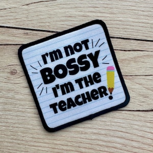 May include: A white patch with black text that reads "I'm not bossy I'm the teacher!" with a yellow pencil with a pink eraser on the right side. The patch has a black border and a white background with blue lines.