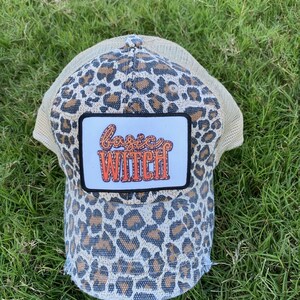 May include: A leopard print trucker hat with a patch that says "basic witch" in orange letters.
