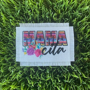 May include: A white canvas with a colorful "MAMA" design in a striped pattern and the word "cita" in black lettering. The design is surrounded by a floral border.