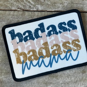 May include: A white patch with black edging features the text "badass mama" in a layered, retro font style. The text is in shades of blue, brown, and pink.