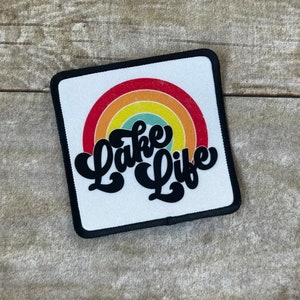 May include: A white patch with a black border featuring a rainbow with a red, orange, yellow, green, and blue color scheme. The text "Lake Life" is written in a black, cursive font.