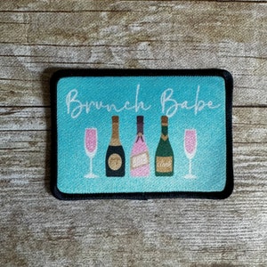 May include: A blue and black rectangular patch with the text "Brunch Babe" in white script. There are four bottles of champagne and two champagne flutes in the center of the patch.