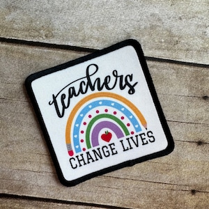 May include: A white square patch with a black border. The patch features a rainbow with a red apple in the center and the text "teachers change lives" in black.
