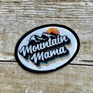 May include: A white oval patch with black stitching around the edges. The patch features a black and white graphic of mountains with a setting sun behind them. The text "Mountain Mama" is written in a retro font with a rainbow-colored design.