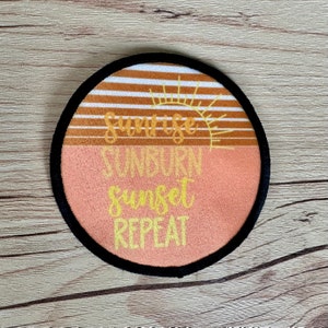 May include: Round patch with a black border and a sunset graphic. The patch has a brown and white striped background with a yellow sun and the text "Sunrise Sunburn Sunset Repeat" in yellow.