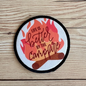 May include: A white round patch with black stitching around the edges. The patch features a red and orange flame design with two brown logs at the bottom. The text "Life is better by the campfire" is written in red cursive.