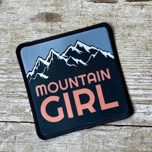 May include: Square patch with a black border featuring a mountain range graphic in white and black against a light blue sky. The words "MOUNTAIN GIRL" are printed in a coral color.