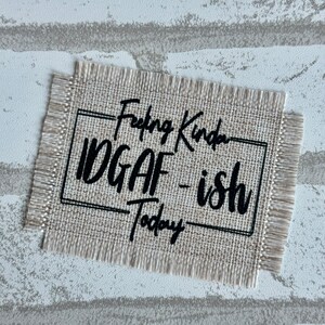 May include: A beige burlap fabric patch with black embroidered text that reads "Feeling Kinda IDGAF - ish Today".