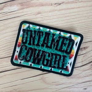 May include: Rectangular patch with the words "UNTAMED COWGIRL" in black, distressed lettering. The background features a turquoise leopard print, bordered by a black edge and a Southwestern-inspired pattern in white, turquoise, orange, and yellow.