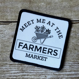 May include: White patch with black text and a black border. The text reads "Meet me at the Farmers Market". A black and white illustration of a wheelbarrow with plants is above the text.