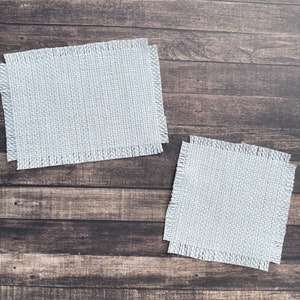 May include: Two white, rectangular, woven placemats with frayed edges on a brown wooden surface.