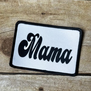 May include: A rectangular patch with the word "Mama" in a black, retro script font. The patch has a white background and a black border. The patch is likely for clothing or accessories.