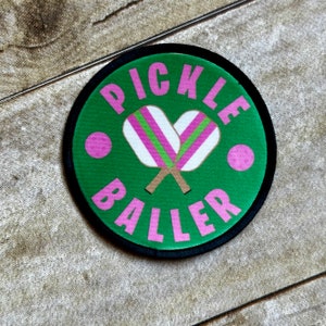 May include: A green and black circular patch with the text "Pickle Baller" in pink lettering. The patch features a pair of pink and green pickleball paddles.